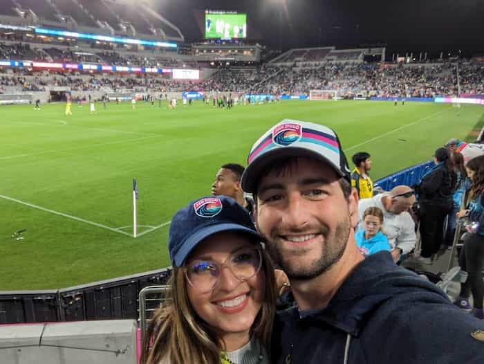 Selfie of a man and women in SD Wave hats with the corner of a soccer field behind them.