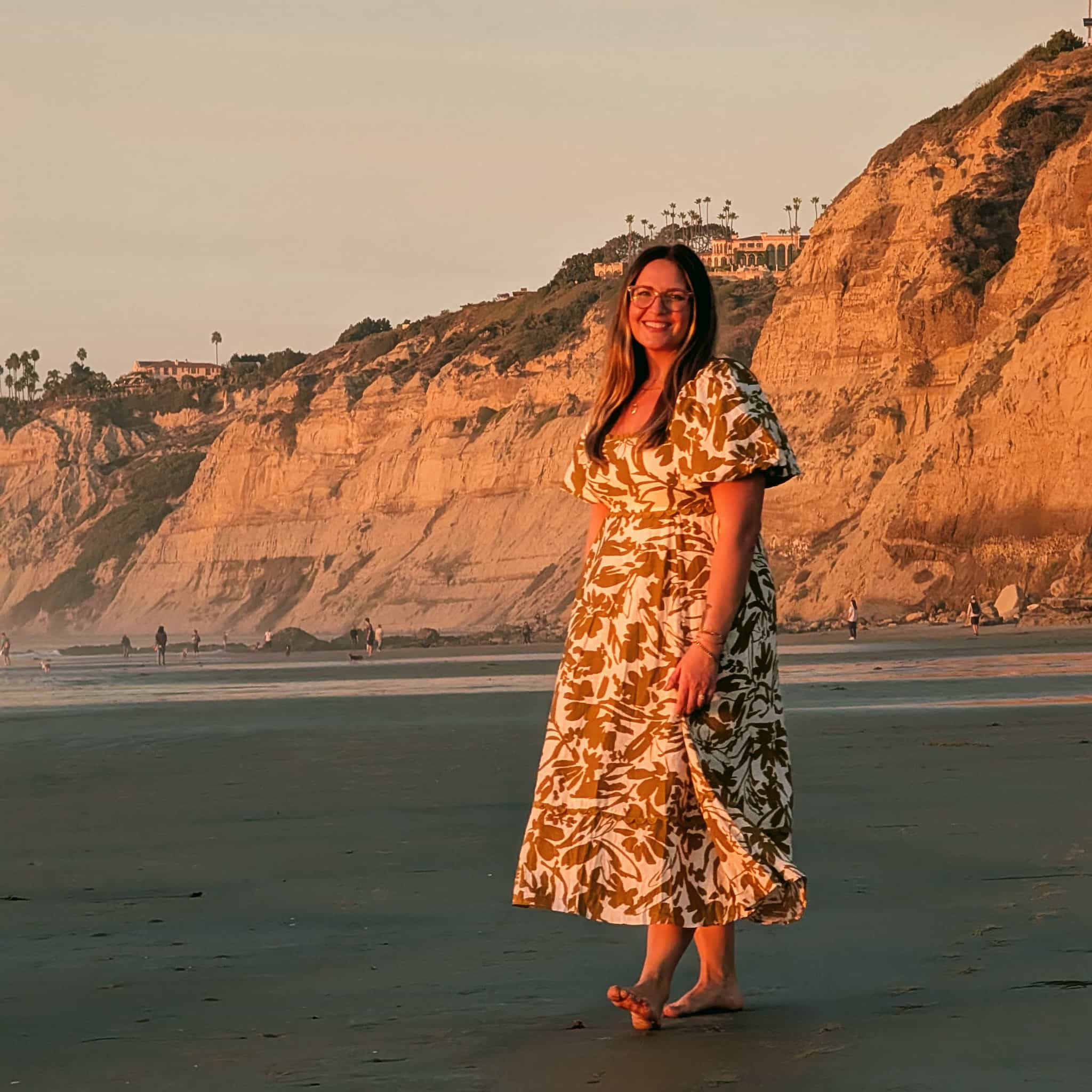 Beautiful woman in a dress posing barefoot on a beach in the warm evening sunlight with large sandy bluffs in the background.