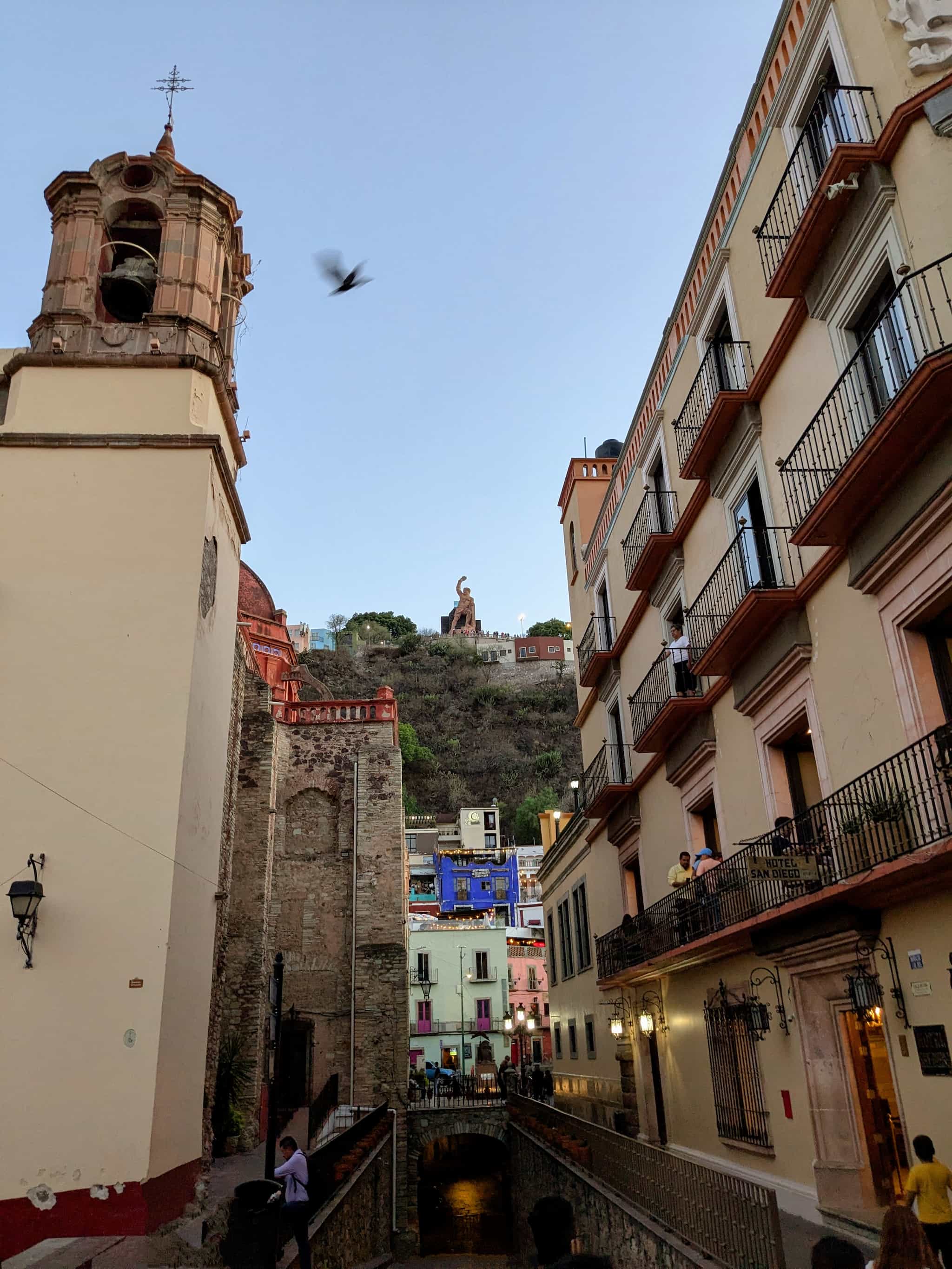 Looking up at a hill with a statue with a tunnel below, church to the side, colorful old buildings, and a flying pigeon.