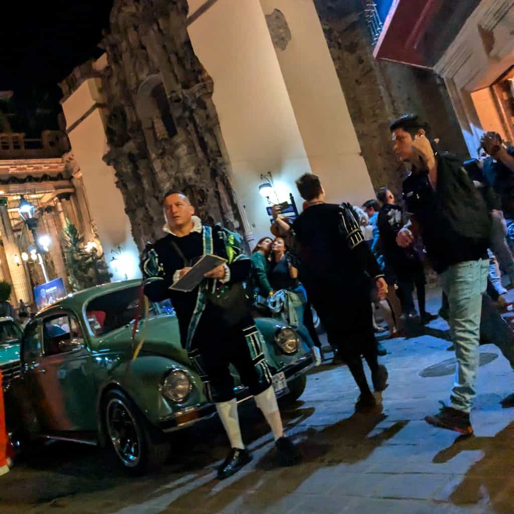 A man dressed in an almost goofy-looking aristocratic garment stands in front of an antique Volkswagon beetle car with lots of people walking around at night between stone building.