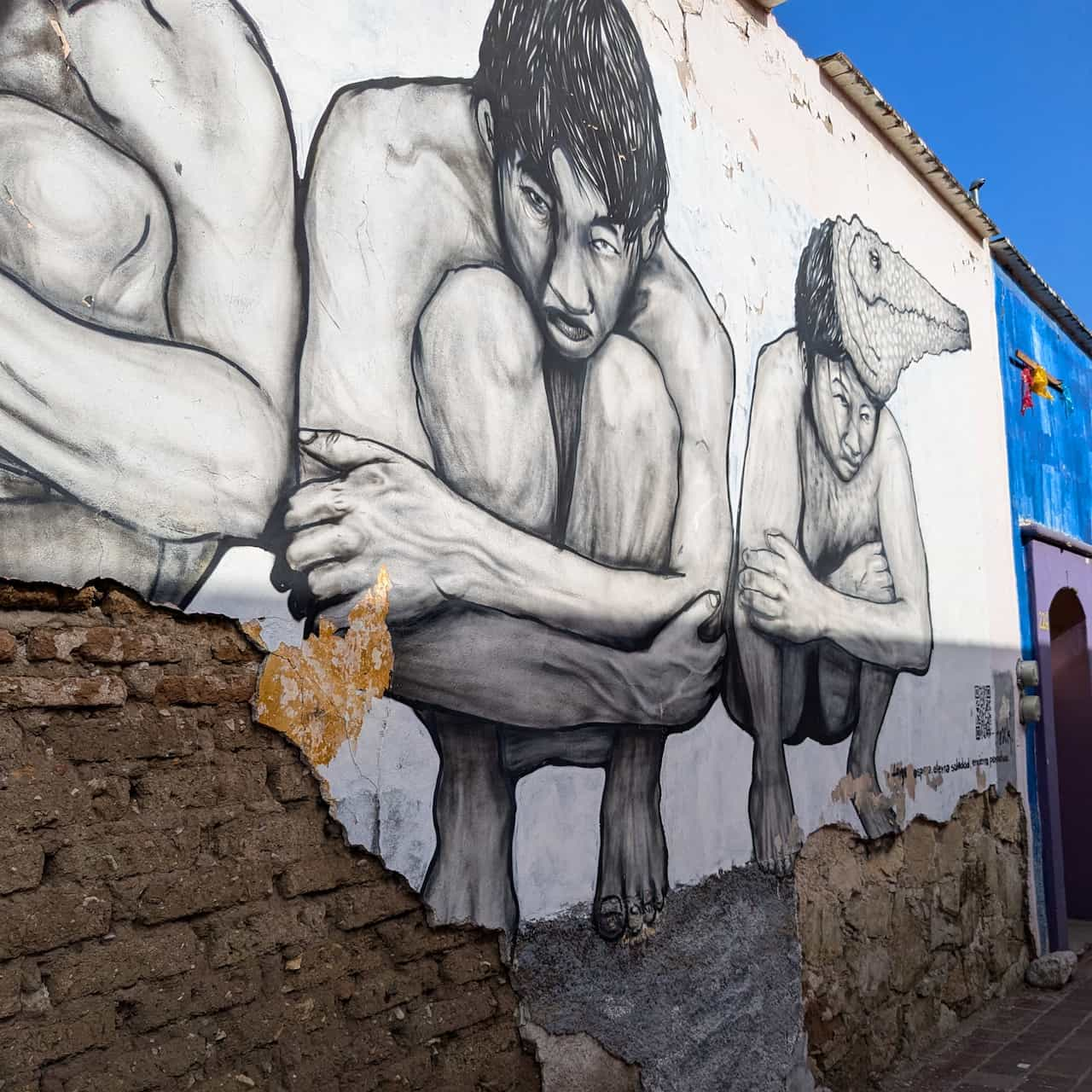 Mural of three men crouched and holding their knees. The bottom of the mural is broken off, revealing the mud bricks of the building's wall.