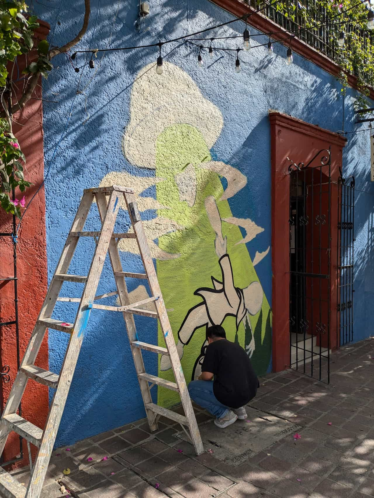 A man with a ladder crouching in front of a partially complete mural of a man being sucked up to a UFO by aliens.