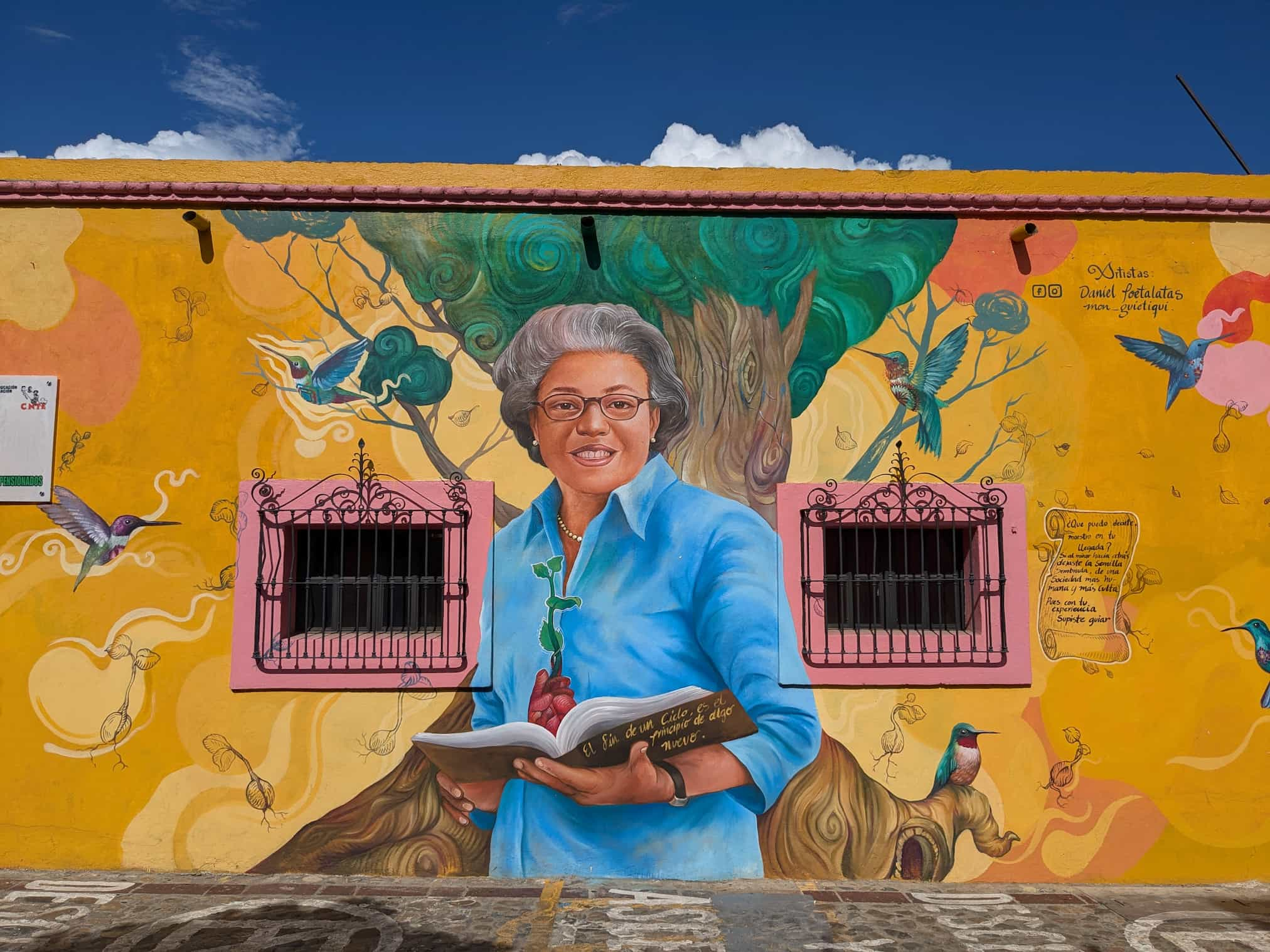 Mural of an older woman standing in front of a tree and holding a book while surrounded by large hummingbirds. The bright color of the mural is bordered by a bright blue sky and cobblestone street.