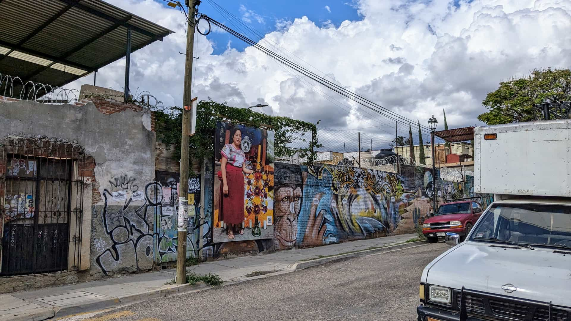 Cement wall topped with barbed wire and painted with murals and graffiti seen from across the street.