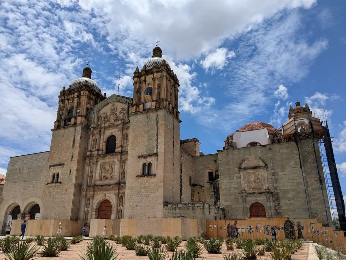 Wider angle view of a large tan stone church in bright light against a blue sky with white clouds.