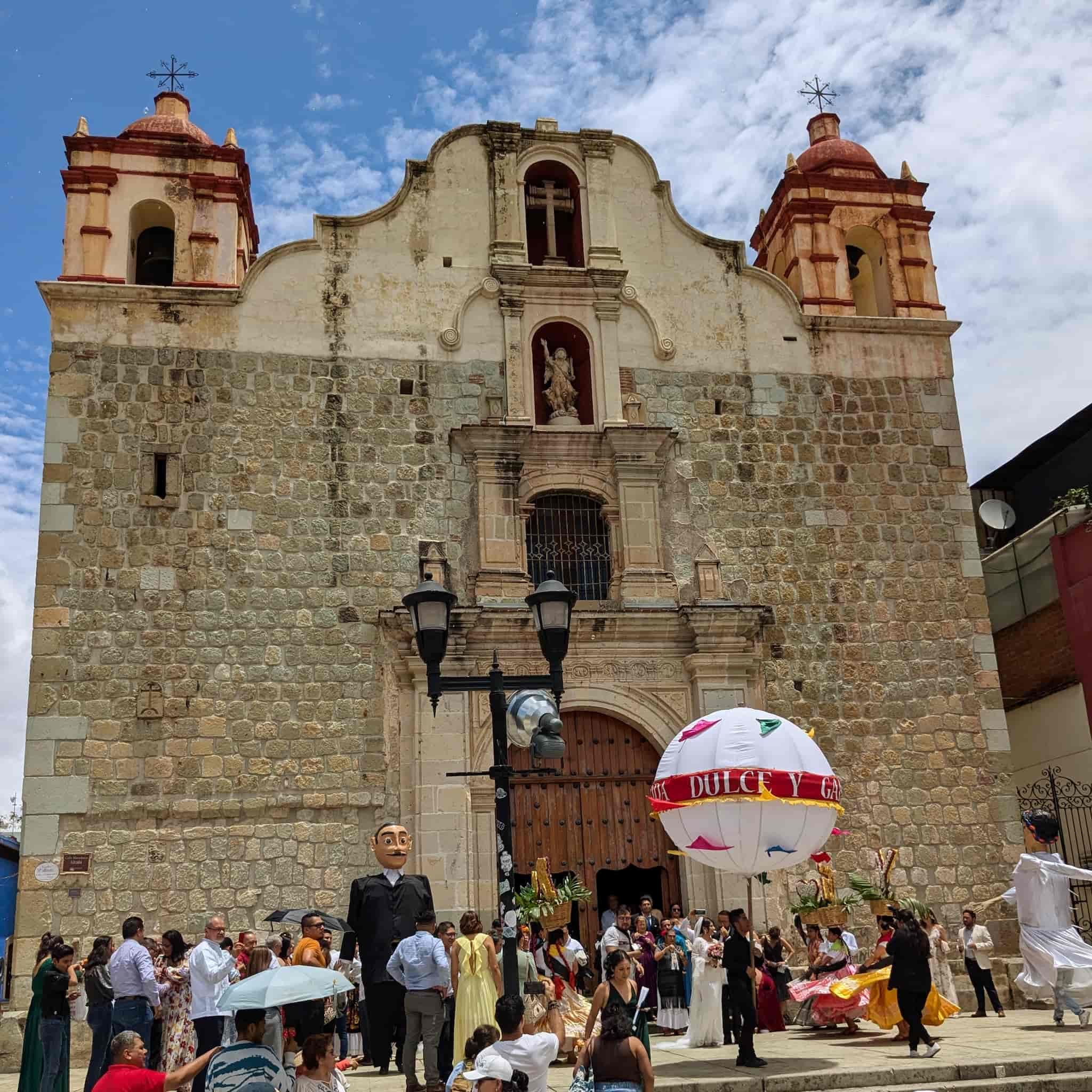 A bunch of people in front of a big stone church with giant floats of a bride and groom.