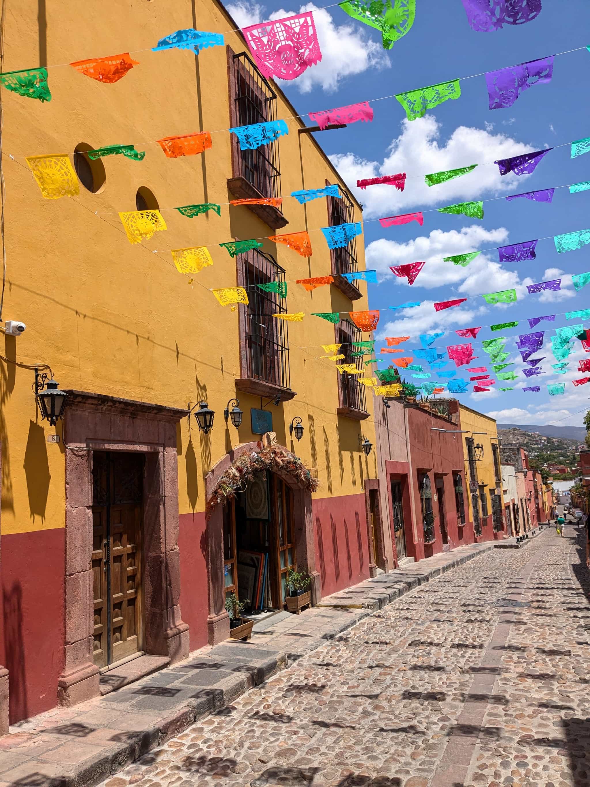 Cobblestone street with yellow buildings and colorful paper flags hanging above on a sunny day.