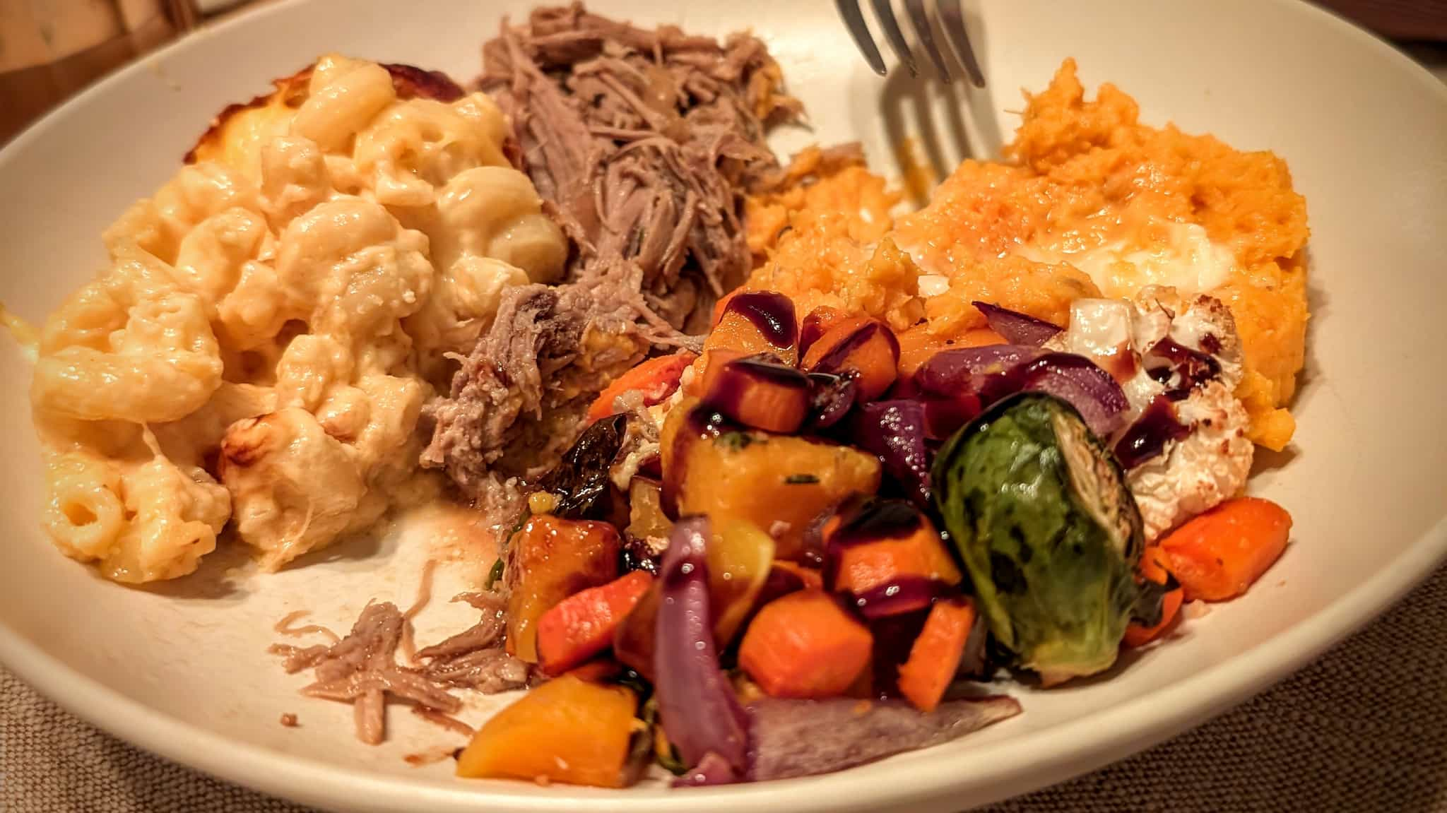 A fork about to dive into a plate full of pulled pork, colorful roasted veggies, mashed sweet potatoes, and mac and cheese in warm lighting.