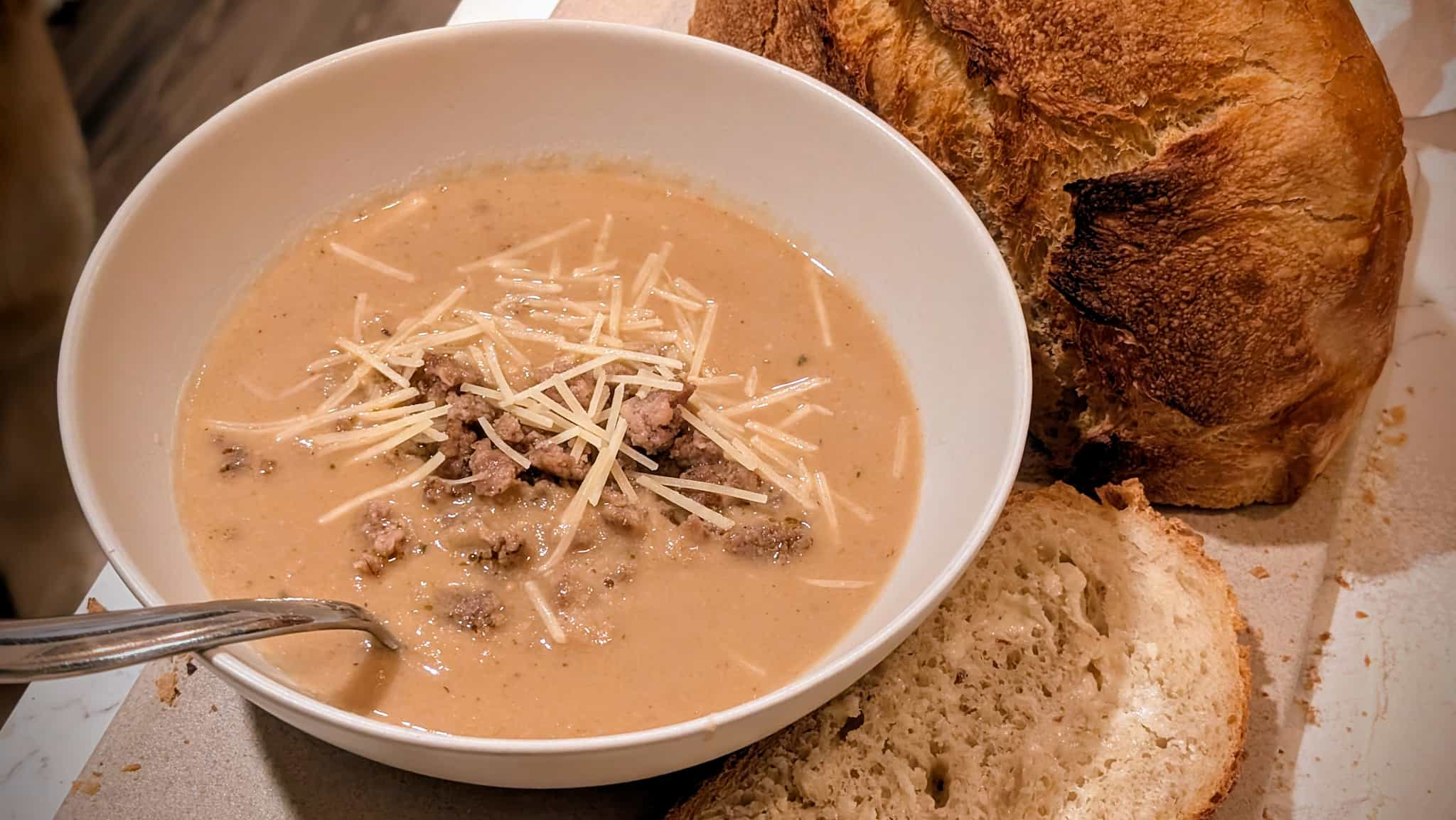 A bowl of white soup with ground sausage and cheese next to bread.