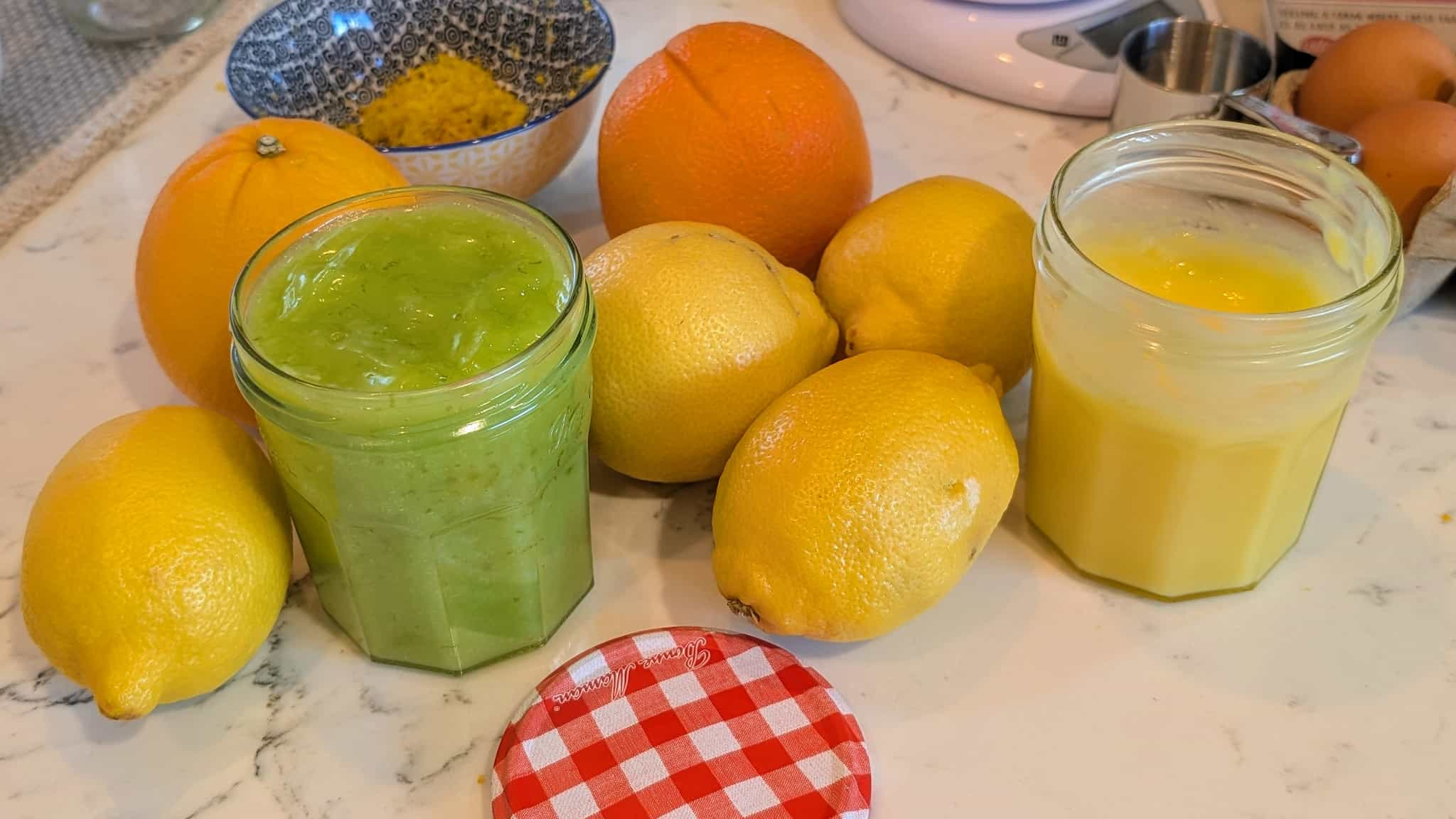 A countertop of lemons and small jars of lemon and lime curd with egs in the background.