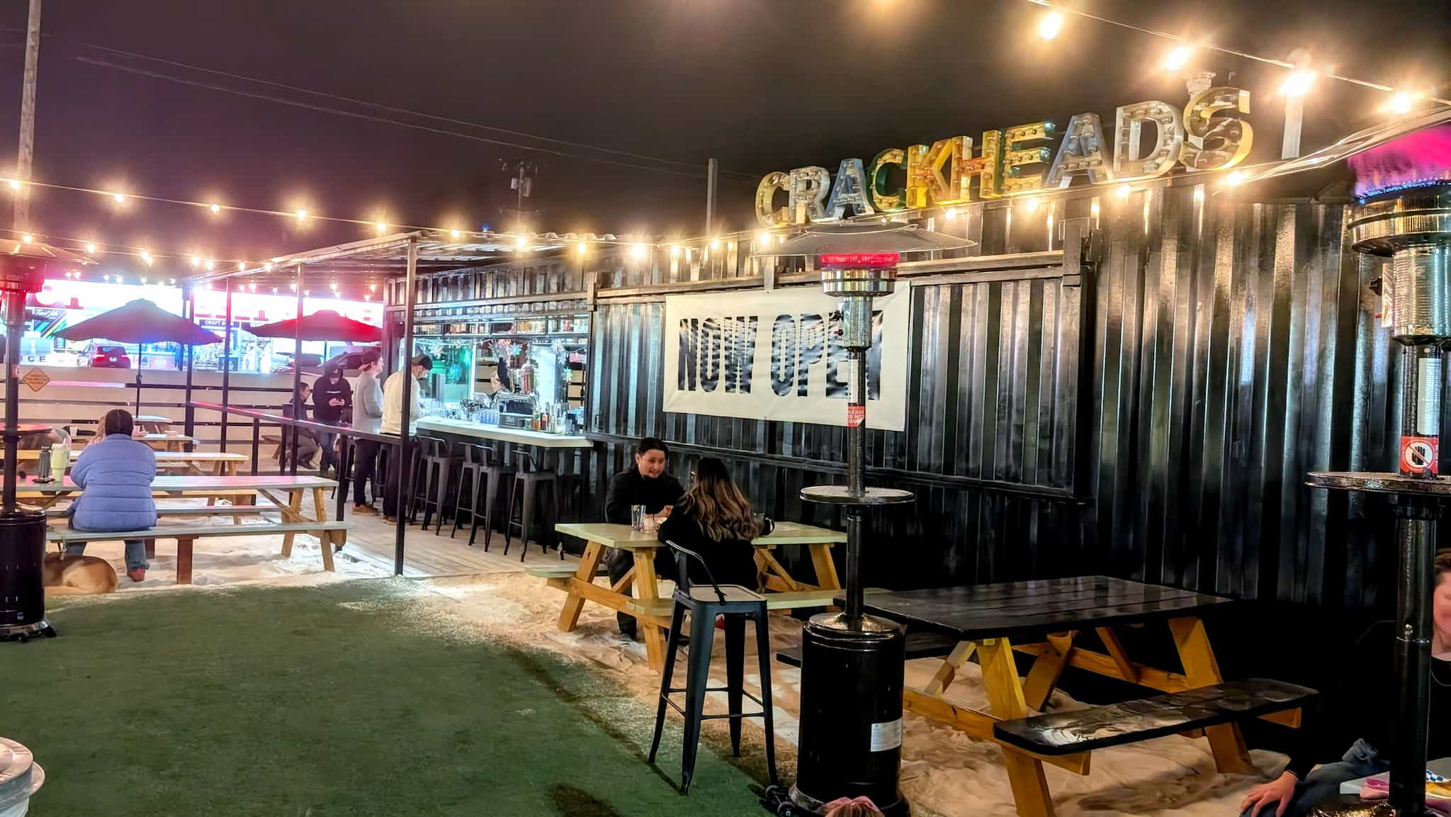 Nighttime photo of a shipping container turned into a bar with picnic tables on sand.