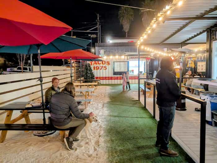 People sitting at picnic tables on sand next to a turf path leading to a shipping container with 'Tacos' written on the side and a serving window.